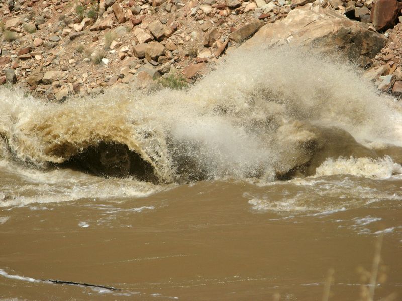 Silt-brown floodwater curls into a breaking wave, its spray bursting against a talus slope of red and tan boulders.