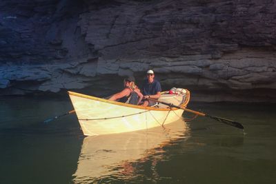 Two people sit in a wooden dory lit gold by low evening sun, drifting on still green water against a shaded, ledged canyon wall.