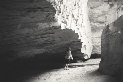Black-and-white view of a lone hiker with a small backpack walking the sandy floor of a desert canyon beneath a huge overhanging sandstone wall, bright light raking the cliff above deep shade.