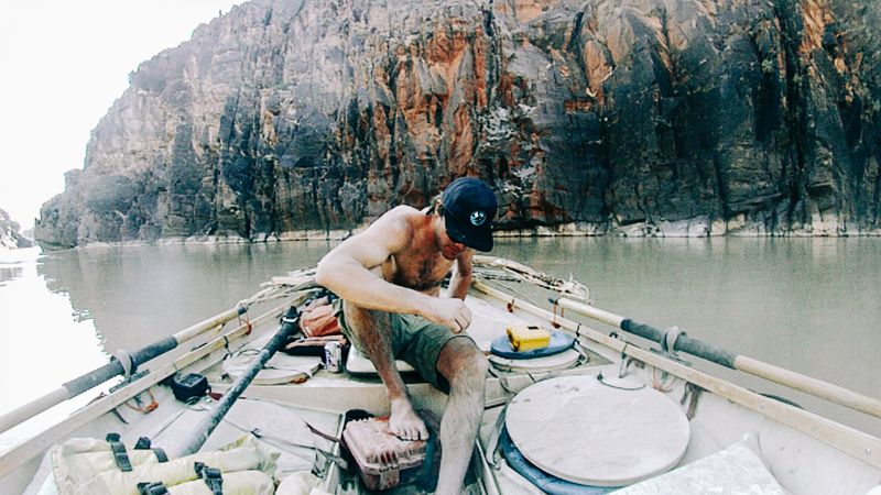 A mud-spattered shirtless boatman in a black cap sits on the front deck of a dory mid-river, bare foot braced on a battered hard case, oars shipped, beneath dark cliffs streaked with orange.