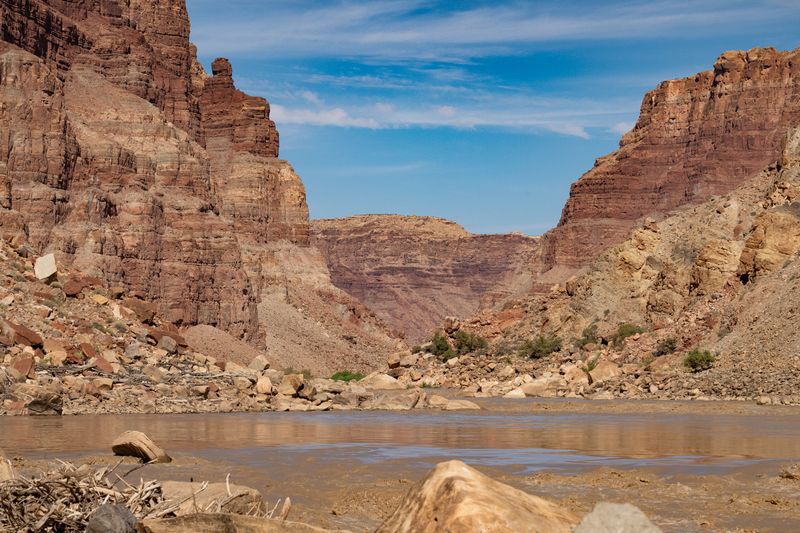 A low view across smooth muddy water and the edge of a rapid toward red layered canyon walls framing a distant mesa