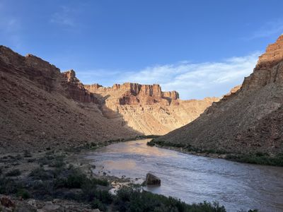 Upstream view of the Colorado River near Range Canyon at evening, the muddy channel curving between shadowed talus slopes toward banded red cliffs holding the last low sunlight.