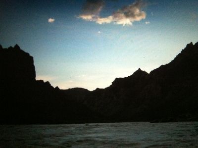Dusk on the water in Cataract Canyon, with canyon rims silhouetted black on both sides, a single pink-lit cloud in a deepening blue sky, and dark rippled current in the foreground.