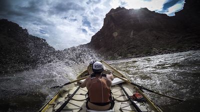 From the stern of a dory entering Big Drop 3, the rower hauls on the oars as whitewater erupts off the bow, with the sun flaring over a dark canyon rim under storm clouds.