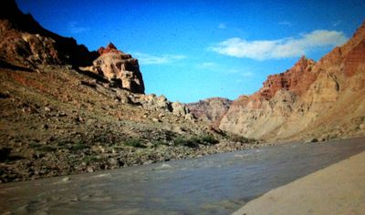 Grainy view from a sandy beach in Cataract Canyon, looking across the silty river to banded red-and-tan canyon walls beneath a deep blue sky.