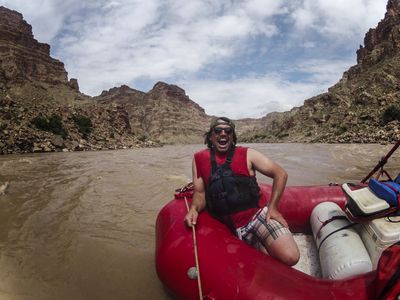 A laughing boatman in sunglasses, backwards cap, and black life vest braces on a gear-loaded red raft mid-river, gripping a line as muddy brown water swirls past beneath cloudy skies.