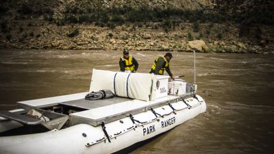 Two National Park Service river rangers in yellow life vests work aboard a white Park Ranger patrol rig on fast, silty high water below a rocky bank in Cataract Canyon.