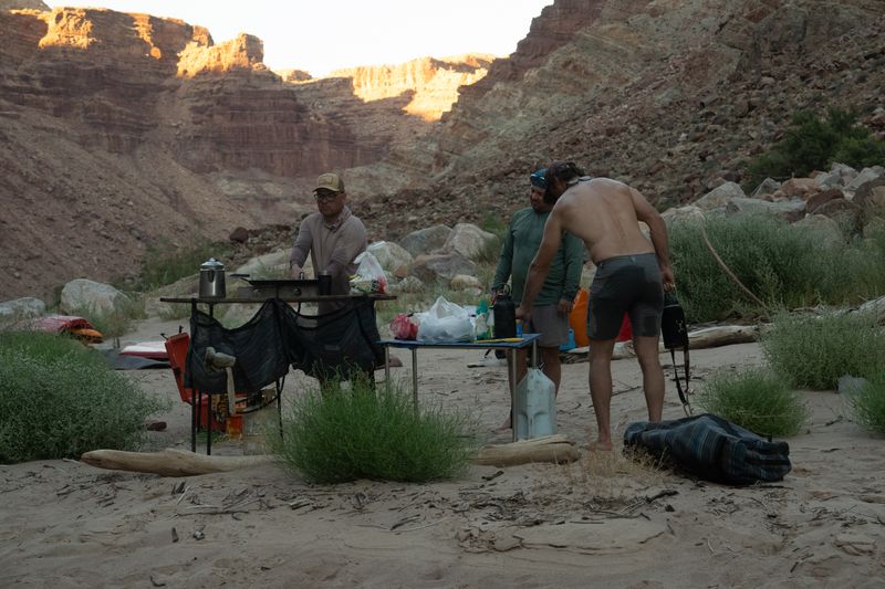 Three men work around folding camp-kitchen tables on a sandy beach, a coffee pot and stove on one, water jugs and food bags on the other