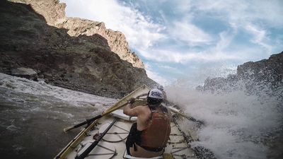 From a dory in Rapid 13 of Cataract Canyon, a wave bursts over the right gunwale as the rower in an orange life vest and white trucker cap pulls on the oars beneath broken canyon slopes.