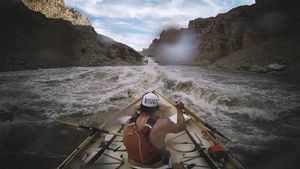 Seen from behind, an oarsman in an orange life vest and white trucker cap rows a white dory into a churning wave train at Big Drop 3, canyon walls half in shadow under a bright, partly cloudy sky.