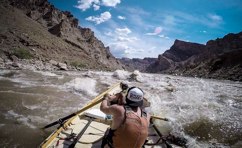From the stern of a dory, an oarsman in an orange life vest and white trucker cap rows toward a muddy wave train in the North Sea rapid of Cataract Canyon, a passenger ducked in the bow and red-brown canyon walls rising under puffy clouds.