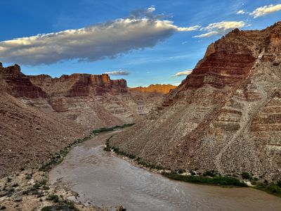 Upstream view of the Colorado River from a bench above Range Creek at evening, the silty river bending beneath banded maroon-and-gray slopes while far cliffs catch orange last light under a blue sky.