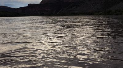Water-level view down the Colorado River at last light, silvered ripples filling most of the frame beneath a dark mesa rim and brush-lined banks.