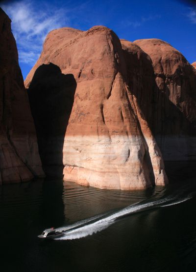 A small powerboat trails a long wake across still dark reservoir water beneath a huge sunlit sandstone dome, a pale bathtub ring marking the old waterline across its base.