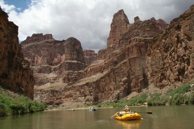 A boater in an orange life jacket rows a yellow raft down calm green water toward towering red pinnacles under building storm clouds.