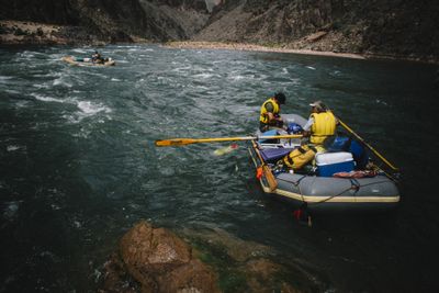 Two boaters in yellow life jackets row a gear-loaded gray raft through choppy green water in a dark rock gorge, with a second oar raft upstream.