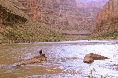 A person sits on a boulder at the edge of the calm, muddy Colorado River beneath tall tiers of layered canyon wall.
