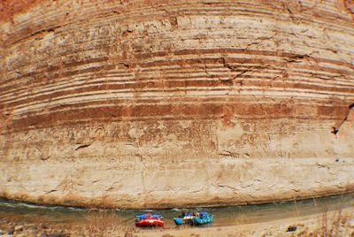 Two loaded rafts sit beached on a narrow shore at the base of a towering rock wall banded with dark and light stripes.
