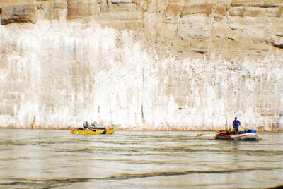 Two small boats drift on flat water beneath a canyon wall streaked with white mineral stains, the scene washed out in bright glare.
