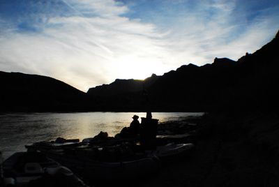 Gear-loaded rafts beached at dusk with a boater in a hat silhouetted against light reflecting off the river.