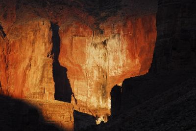 A towering sandstone canyon wall in the Grand Canyon, raked by low-angle warm light that isolates a slender rock pinnacle in silhouette against the glowing cliff face while the foreground talus falls into deep shadow.