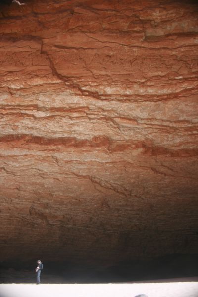 A lone person stands on sand in deep shade at the base of an immense red-and-white banded cliff.
