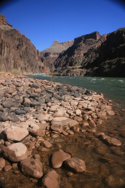 Wet cobbles line the shore of the green Colorado River where a small rapid breaks beneath dark schist cliffs under a deep blue sky.