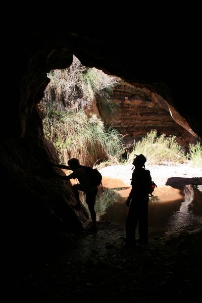 Two hikers stand silhouetted inside a dark grotto, looking out toward a sunlit pool and green brush in a side canyon.