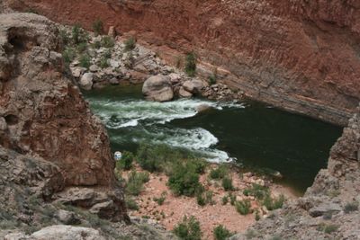 A small rapid breaks across the dark green river between red walls, with a small boat pulled in among rocks on the near shore.