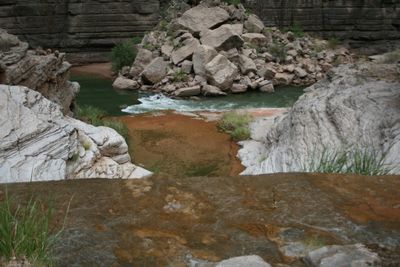 Green water pushes through a boulder pile past a wet orange mineral shelf below dark, banded canyon walls.