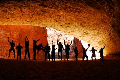 A river crew jumps and poses in silhouette on the sand floor of Redwall Cavern, beneath its huge glowing rock arch above the Colorado River.