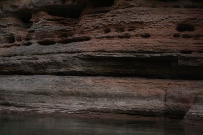 Dark layered rock ledges pocked with small cavities rise from quiet green water in low light.