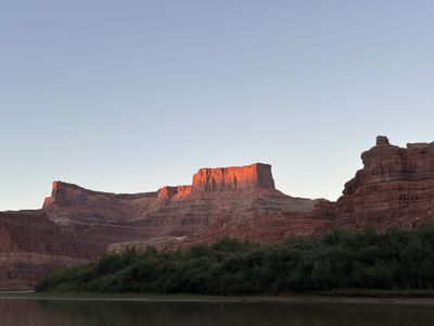 Sunset alpenglow on a distant flat-topped mesa above Meander Canyon, with dense green willows lining the Colorado River bank and the water lying in shadow.