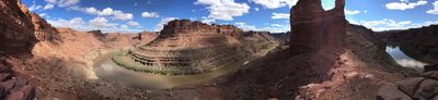 Panorama from the slickrock saddle of the Loop Trail, with the Colorado River bending around both sides of the narrow neck of the meander below broken red canyon walls and scattered clouds.