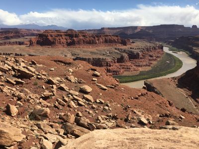 View from a bouldery overlook on the Shafer Trail bench, with the Colorado River curving through a green-lined gooseneck far below and the La Sal Mountains on the horizon.