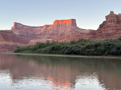 Last light burning orange on a flat-topped mesa high above Meander Canyon, its reflection rippling in the silty Colorado River below a wide green bank.