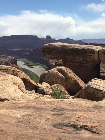 View across bare slickrock and perched boulders from a rim above Meander Canyon, with the green-banked Colorado River winding between layered red walls in the distance.