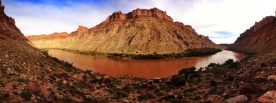 Panorama from a camp bench above a broad bend of the sediment-red Colorado River in Meander Canyon, with a tent pitched among boulders below sunlit canyon walls and a clearing sky.