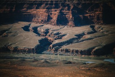 Telephoto winter view from a high overlook, looking down on sage flats, eroded benches, and a drainage cutting toward the green Colorado River beneath shadowed red cliffs in Meander Canyon.