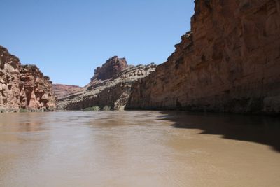 Flat, silty spring water of the Colorado River filling the canyon between a shaded sandstone wall and pocketed red cliffs in The Loop stretch of Meander Canyon.