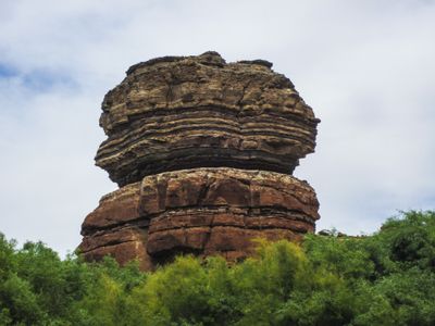 The Coffee Pot, a caprock-topped sandstone tower with thin banded ledges, rising above a screen of green tamarisk on the bank of the Colorado River in Meander Canyon.