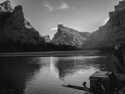 Black-and-white river-level view approaching the confluence of the Green and Colorado rivers, with a crew member in a horned helmet seated by a dry box and oar while sunlight breaks across the canyon walls ahead.
