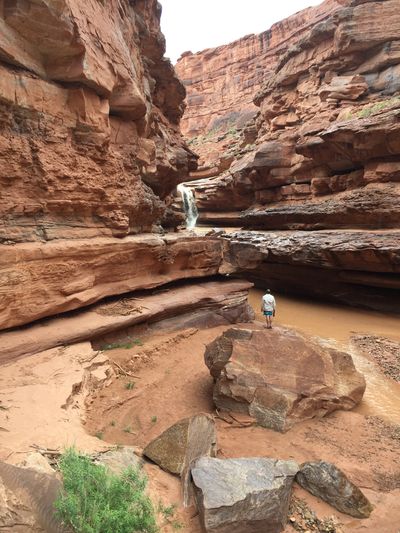 A hiker stands in the muddy pool below a flash-flood waterfall pouring through a slot in the red sandstone cliffs of Indian Creek, a side canyon of Meander Canyon.