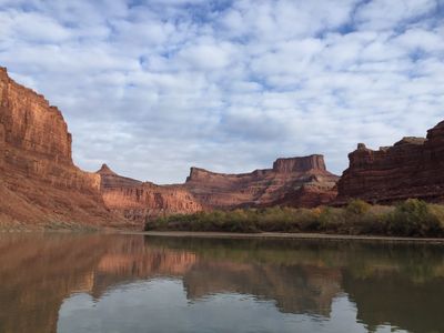 Calm December water in Meander Canyon reflecting red canyon walls, a pointed spire, and the tawny golds of late-season willows under a mackerel sky.
