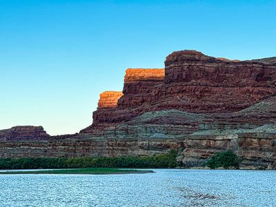 Dusk light banding the caprock of stepped red buttes above the Colorado River in Meander Canyon, with deep-blue rippled water running past a strip of green bank.