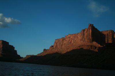 Late sun on a red cliff band and caprock summit above a hard shadow line, with dark rippled water of the Colorado River in the foreground of Meander Canyon.