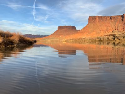 Rippled, glassy water on the Colorado River reflecting orange sandstone cliffs and a butte at golden hour, with dormant tan brush on the far bank.