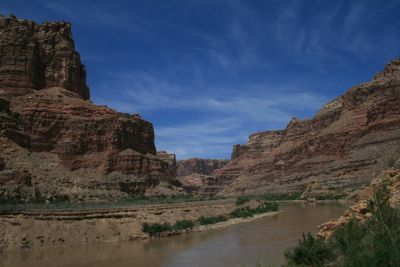 Silt-brown Colorado River curving between layered red and tan sandstone walls in Narrow Canyon under a deep blue sky, with green brush on the low benches.