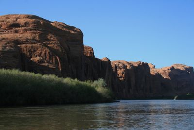 Calm green water on the Colorado River below Moab, with a sunlit band of willows and tamarisk beneath massive shaded sandstone walls in the Portal corridor.