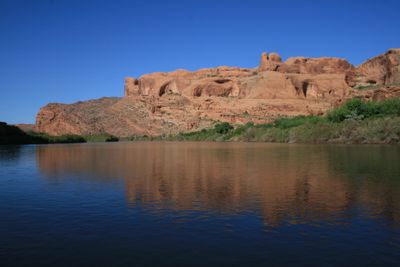 Two-tone river surface on the Colorado below Moab reflecting a red sandstone rim pocked with alcoves, with spring-green cottonwoods on the right bank under a deep blue sky.